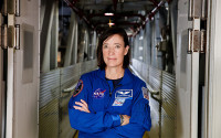 Portarit of a female astronaut with short brown hair wearing a blue jumpsuit. She is standing in a training facility and her arms are crossed.