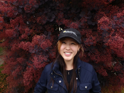 A smiling woman in a baseball cap stands in front of a large plant with pink flowers