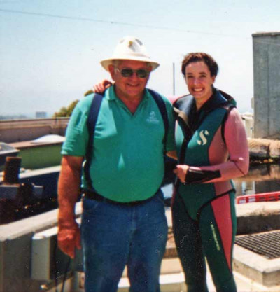 A woman in a diving suit stands next to a man in a green shirt and sun hat.