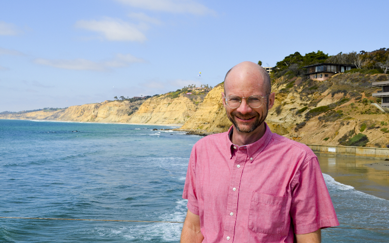 A smiling man in front of the beach with cliffs in the background.