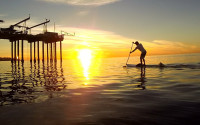 A stand-up paddleboarder near a pier at sunset.