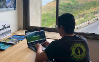A young man works on a laptop in an office overlooking a verdant canyon and the ocean.