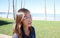 A brunette woman wearing glasses stands near a grassy patch along the coast