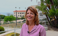 Portrait of a smiling woman wearing a purple shirt; a building and the ocean are seen in the background