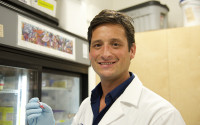 Portrait of a male scientist in a lab; he's wearing a white lab coat and blue gloves