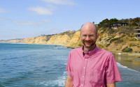 A smiling man in front of the beach with cliffs in the background.