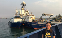 A young woman in a UCSD t-shirt stands in front of a docked ship.