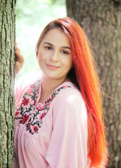 A woman with long orange hair leans against a tree