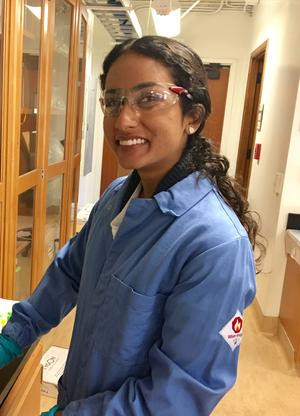 A woman wearing a blue lab coat and clear protective glasses works in a lab