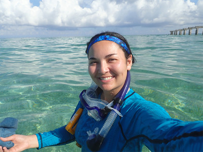 A selfie of a woman wearing a blue rashguard and snorkel gear. She is at a tropical beach and the water is crystal clear.