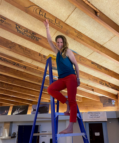A woman on a ladder points to a wooden beam where she signed her name