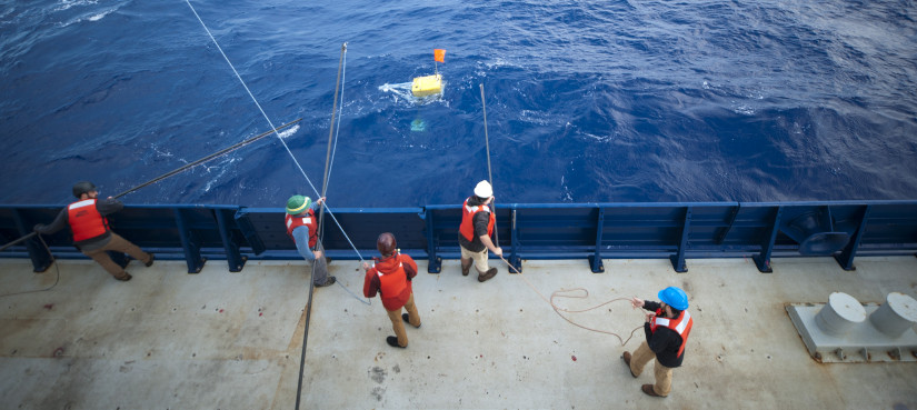A group of researchers out at sea wearing life vests and hard hats, holding large poles stretched over the boat rail into the water.