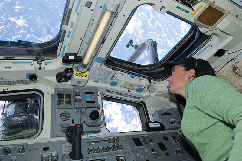 A female astronaut in a space shuttle gazes out the window at Earth.