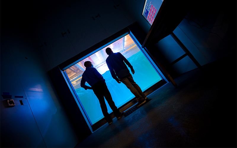 Two men gaze into a wave channel through an observation window.