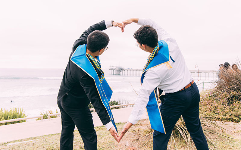 Two graduates holding arms to form heart shape in front of ocean view.